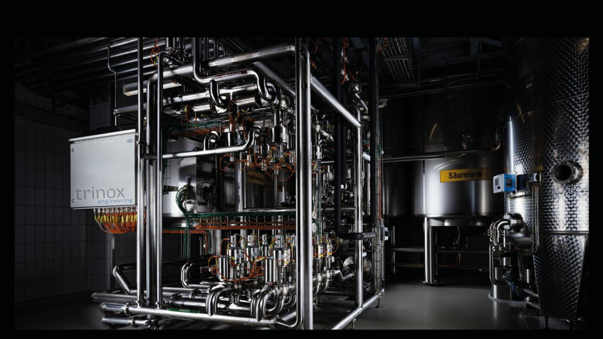 A complex industrial setup with stainless steel pipes, tanks, and a control box labeled trinox in a dark, modern facility; one large tank is labeled Sauerlank.