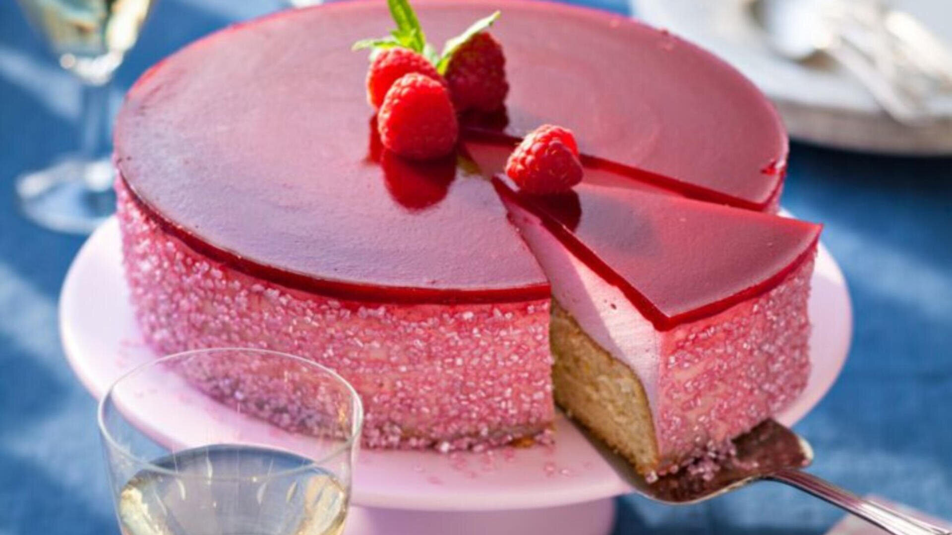 Eine runde Himbeermousse-Torte mit einer glänzenden roten Oberfläche, dekoriert mit frischen Himbeeren und Minze. Es wird ein Stück serviert, das ein helles, cremiges Inneres und einen rosafarbenen, mit Zucker überzogenen äußeren Rand zeigt.