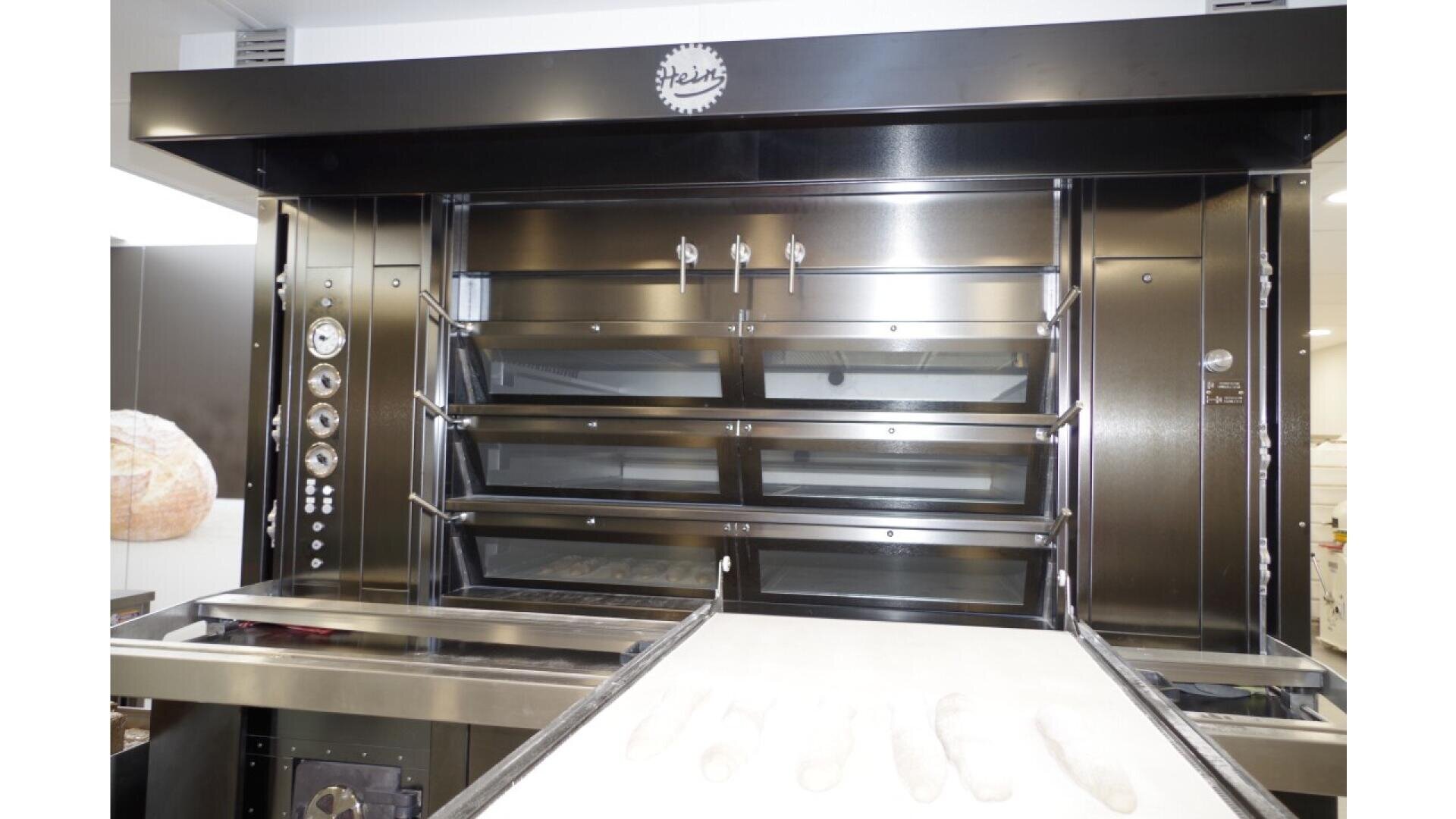 Large commercial bread oven with multiple stacked compartments and control dials on the side; a conveyor belt in front holds several pieces of unbaked dough ready to be loaded into the oven.