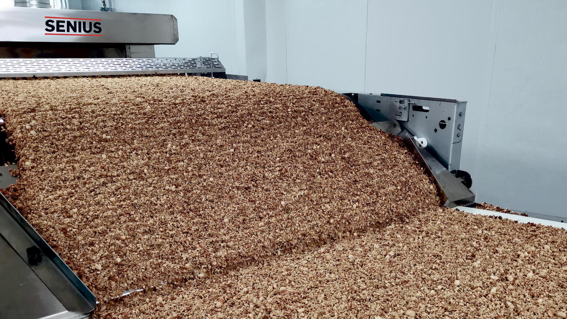 Eine große Industriemaschine mit der Bezeichnung SENIUS verarbeitet und verteilt eine dicke Schicht braunes Müsli oder Getreide auf einem Förderband in einer Lebensmittelproduktionsanlage.