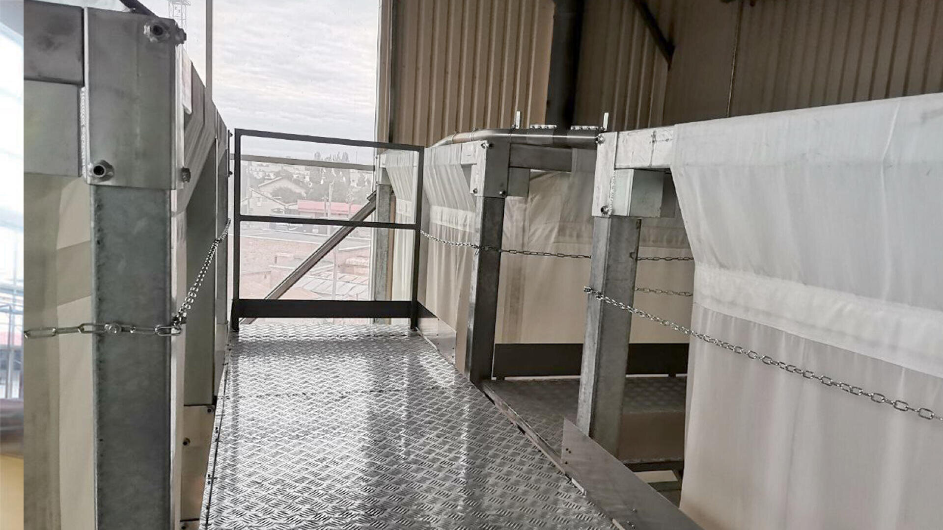 A metal walkway with a diamond plate floor bordered by chains, situated between large industrial tanks or containers, inside a building with windows and corrugated walls.