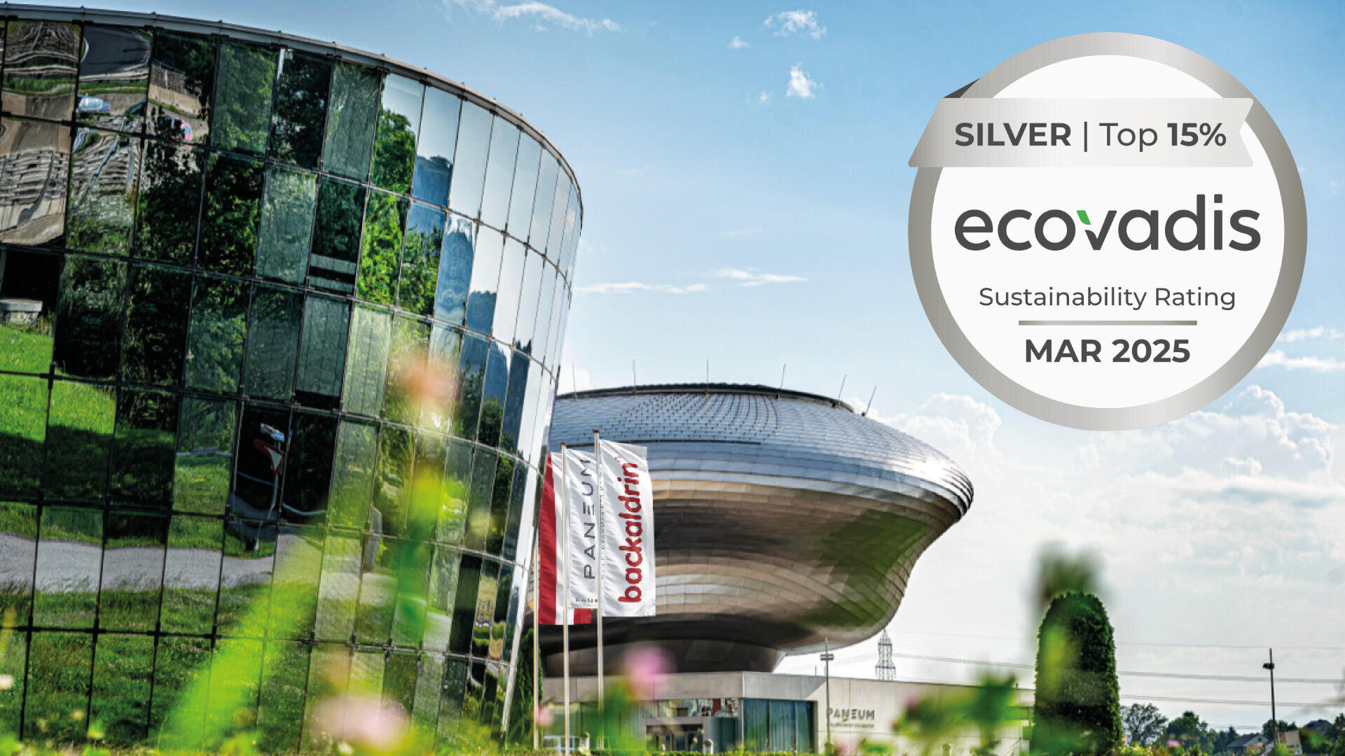 Modernes Glasgebäude und eine futuristische silberne Struktur unter blauem Himmel, davor Fahnen und Grünpflanzen. Ein EcoVadis-Abzeichen zeigt „SILBER | Top 15 %, Nachhaltigkeitsrating, MÄRZ 2025.“.