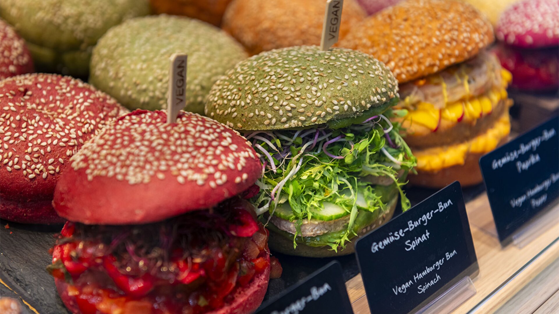 Bunte vegane Burger mit roten, grünen und gelben Sesambrötchen sind in einer Reihe aufgestellt. Jeder Burger ist mit frischem Gemüse und Grünzeug gefüllt und mit kleinen Schildern und "vegan"-Flaggen gekennzeichnet.