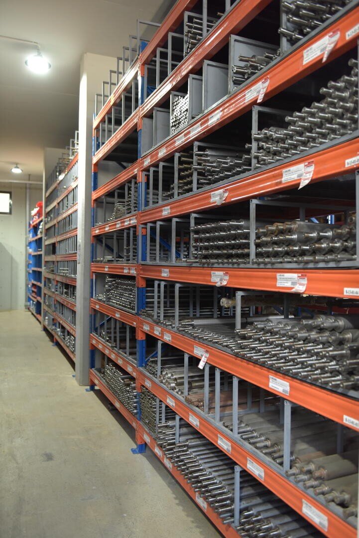 Rows of metal shelves in a warehouse, filled with organized metal rods or parts. The shelves are labeled, and the floor is clean, creating an orderly industrial storage space.