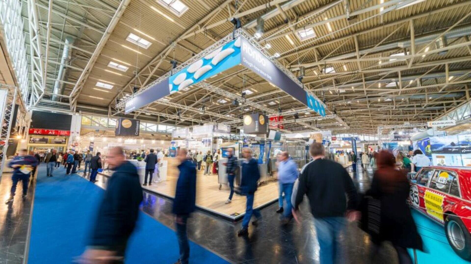 A busy exhibition hall with people walking around various booths and displays under bright overhead lights, featuring a blue carpeted walkway and industrial-style ceiling.
