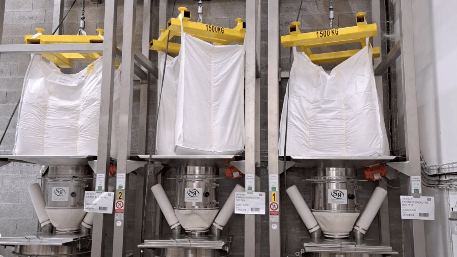 Three large white industrial bags, each labeled 1500 KG, are suspended over stainless steel hoppers in a processing facility, with visible equipment labels and safety signs on the metal frames.