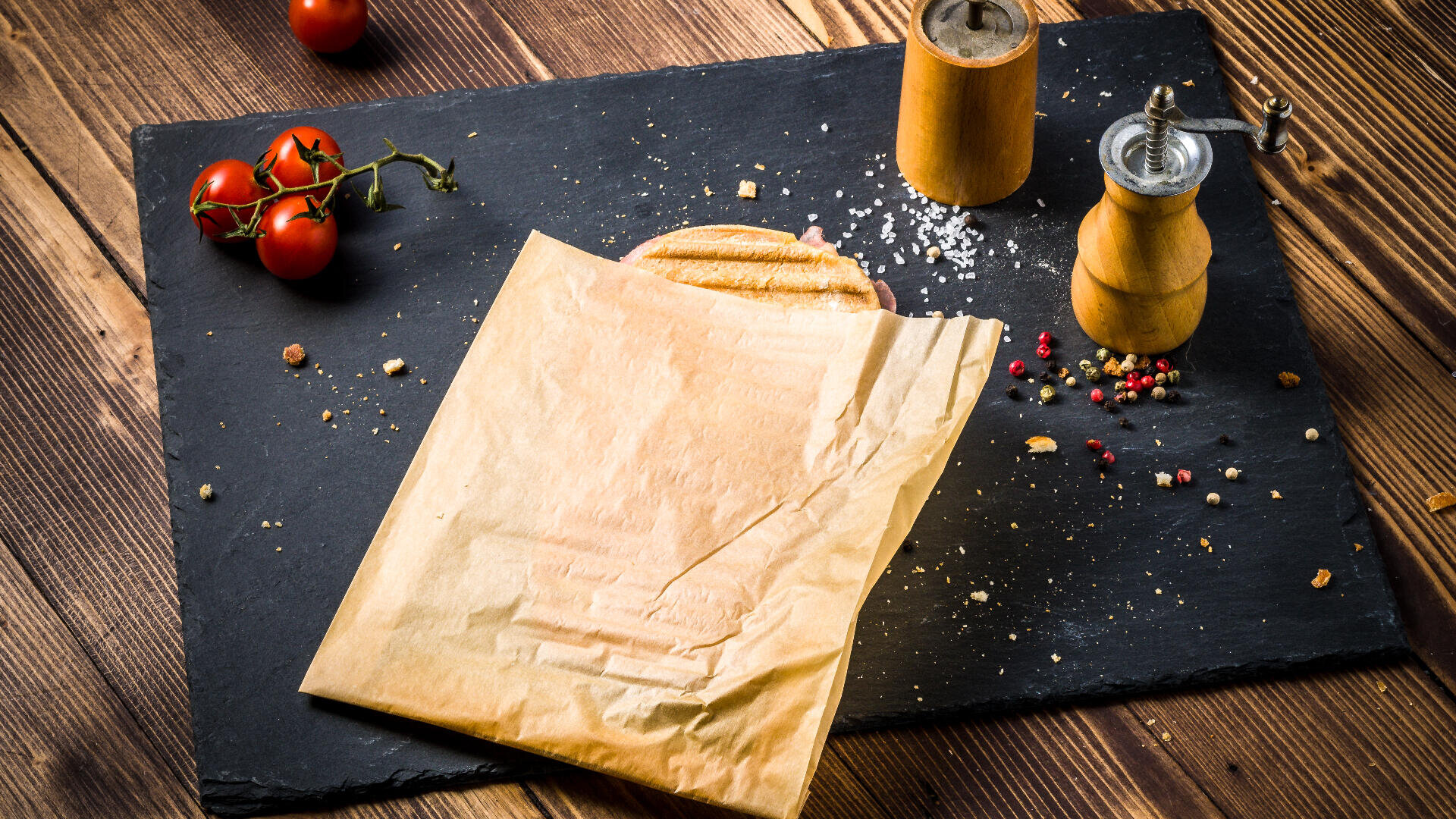 A pita bread partially wrapped in brown paper sits on a black slate board with scattered salt, pepper, a pepper mill, a salt shaker, and vine tomatoes on a rustic wooden table.