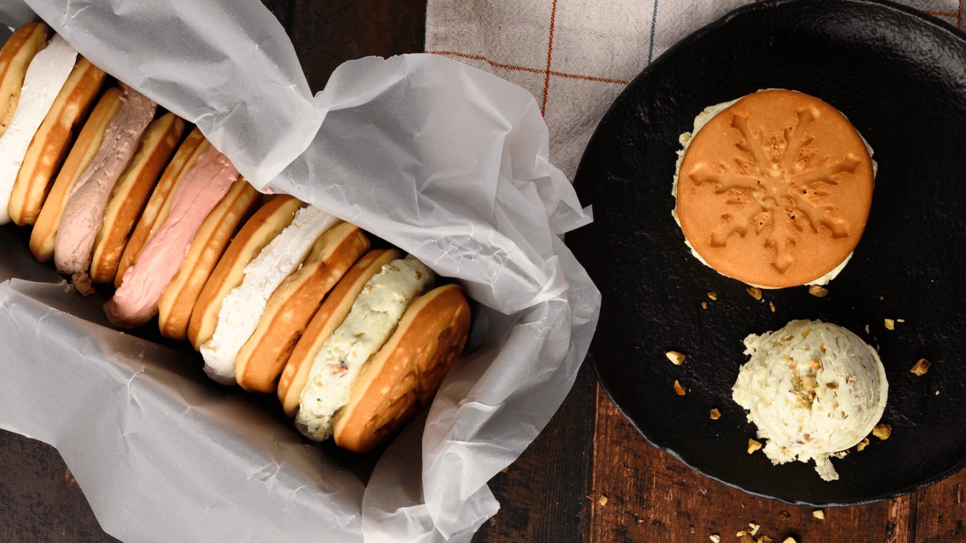A container lined with parchment paper holds several assorted ice cream sandwiches. Next to it, a black plate displays one ice cream sandwich and a scoop of ice cream with scattered nuts on a dark wooden surface.
