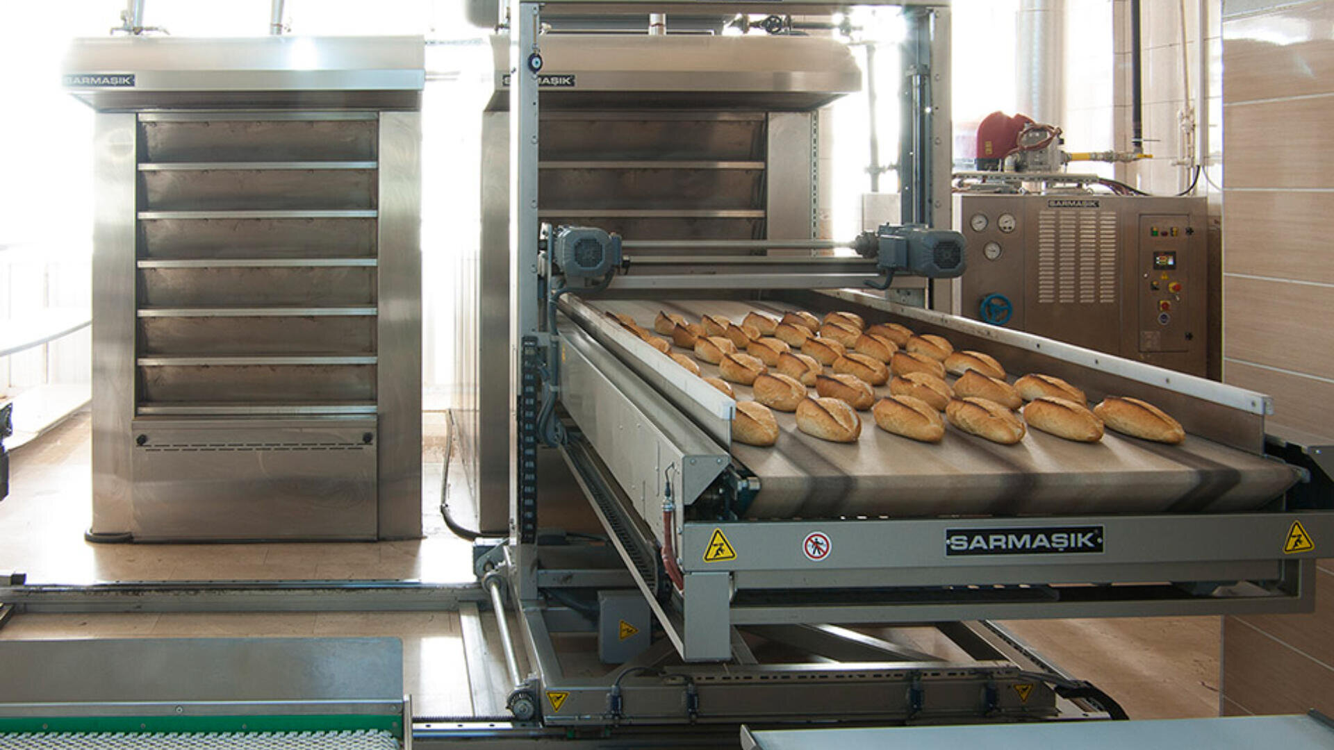 Reihen von frisch gebackenen Brötchen auf einem Förderband in einer industriellen Bäckerei, mit großen Edelstahlöfen und Schalttafeln im Hintergrund.