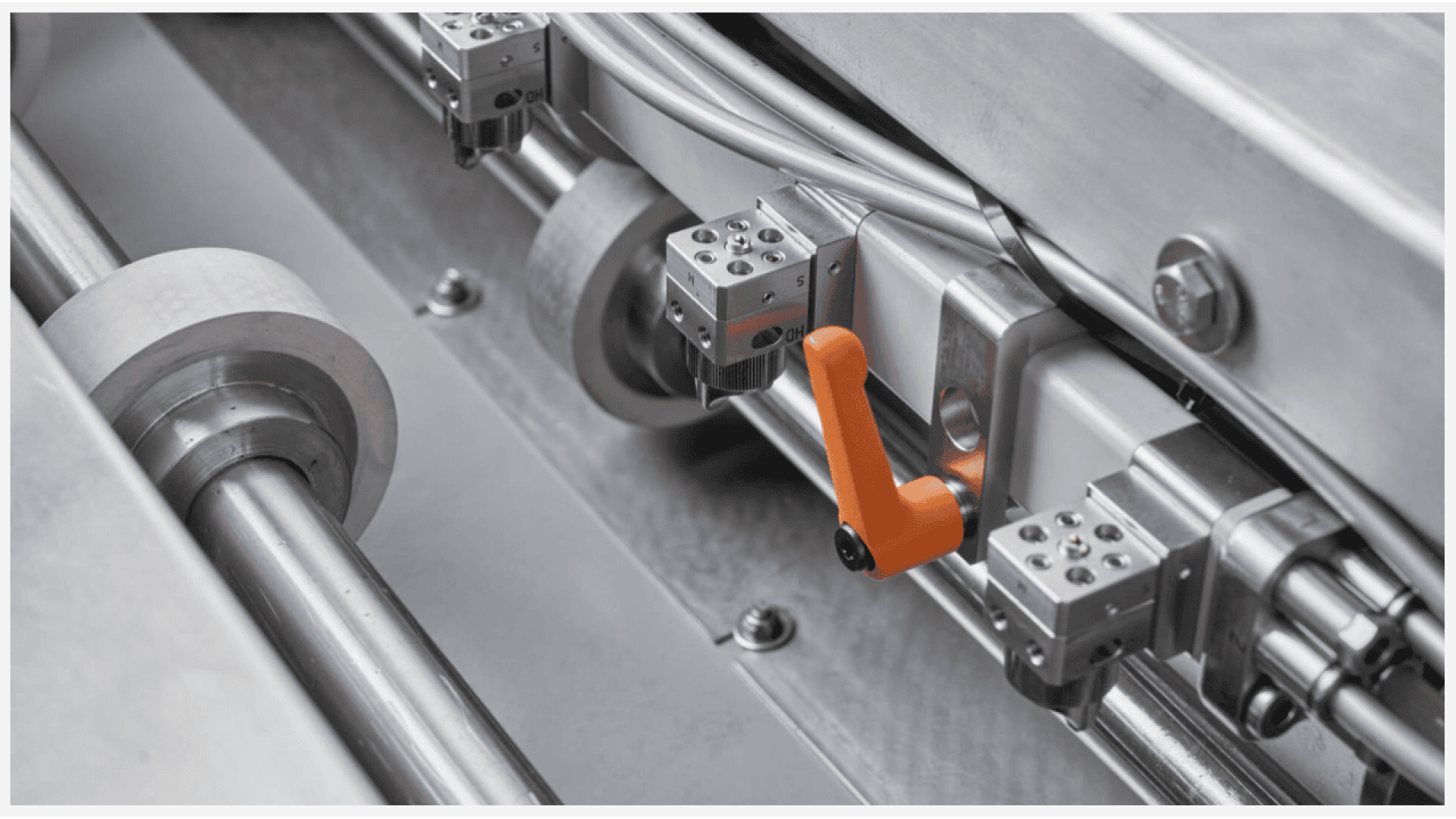 Close-up of a metallic industrial machine with rollers, rails, and components, featuring an orange lever handle attached to a fixture among gray and silver parts.
