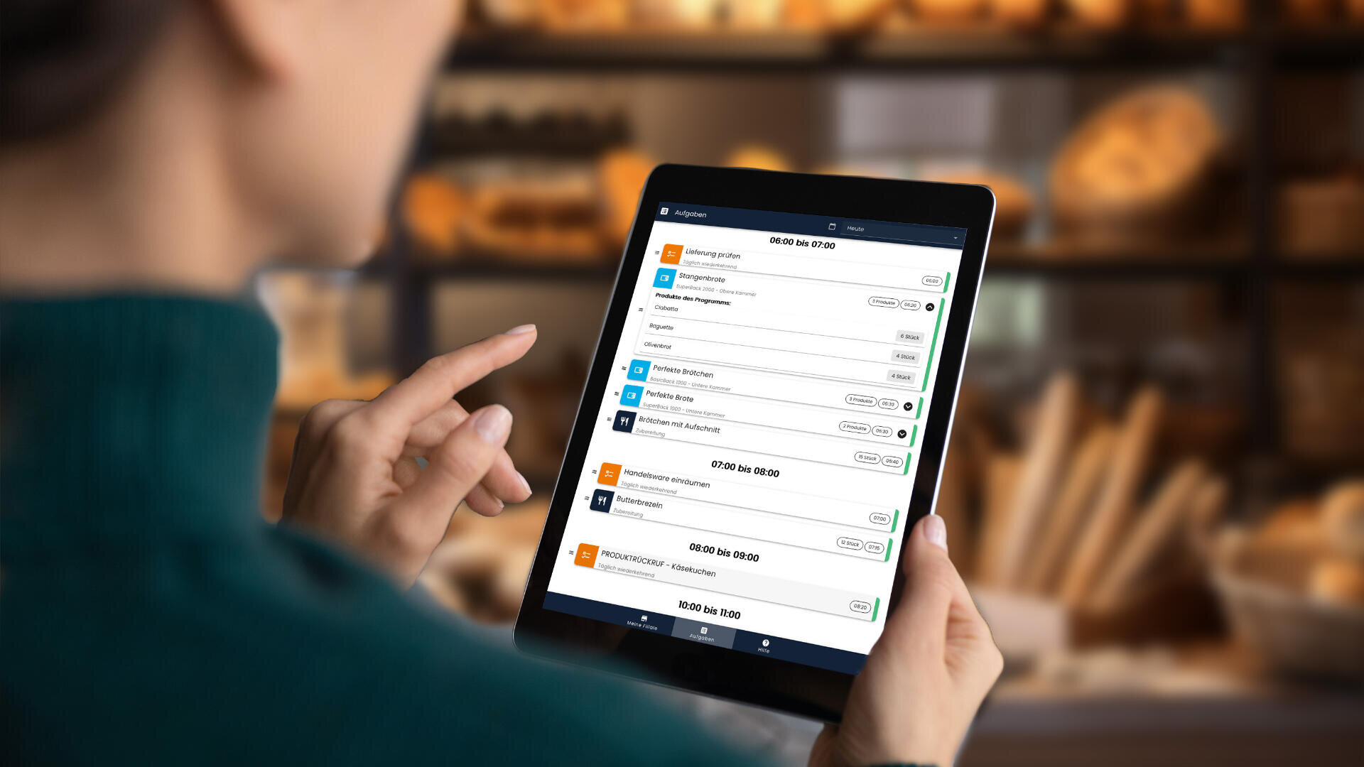 Eine Person hält und benutzt ein Tablet, auf dem eine Terminplanungs- oder Buchungs-App mit einer Liste von Veranstaltungen oder Terminen angezeigt wird. Der Hintergrund ist unscharf und zeigt Regale mit Brot und Gebäck in einer Bäckerei.