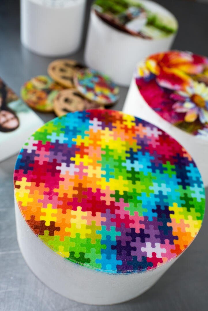 A round box with a lid decorated in a vibrant puzzle piece pattern sits on a table, surrounded by other round boxes and objects with colorful designs and photo prints.