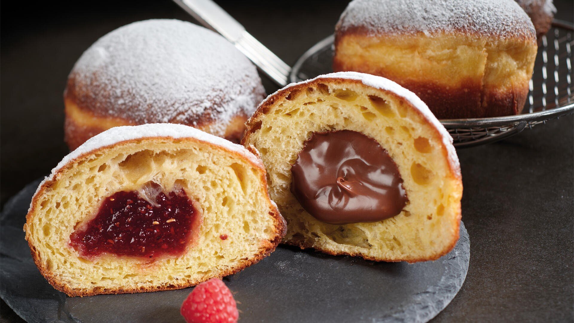Two round doughnuts dusted with powdered sugar, one of which is cut open and contains a chocolate and a raspberry jam filling, lie on a slate tray with a fresh raspberry in front of it.