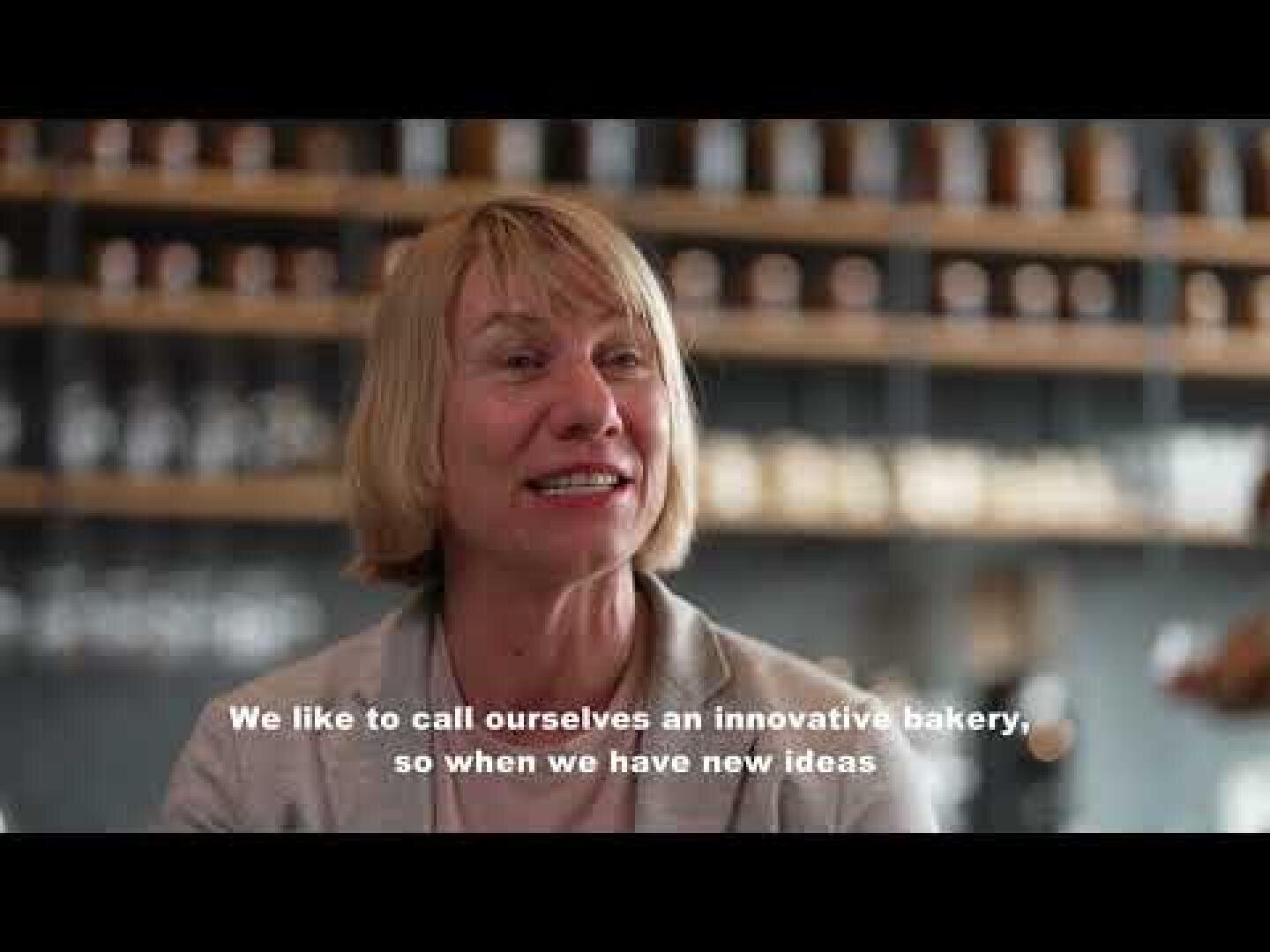 A smiling woman with short blonde hair sits in a modern bakery, shelves with jars in the background. White subtitles read: We like to call ourselves an innovative bakery, so when we have new ideas.