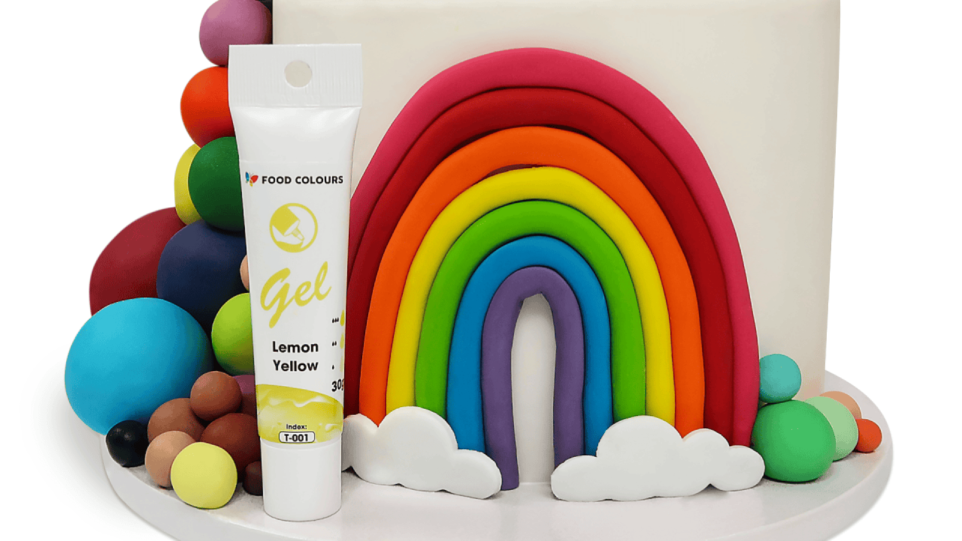 Ein weißer Kuchen, dekoriert mit einem bunten Fondant-Regenbogen, Wolken und Kugeln, mit einer Tube zitronengelber Lebensmittelfarbe davor.