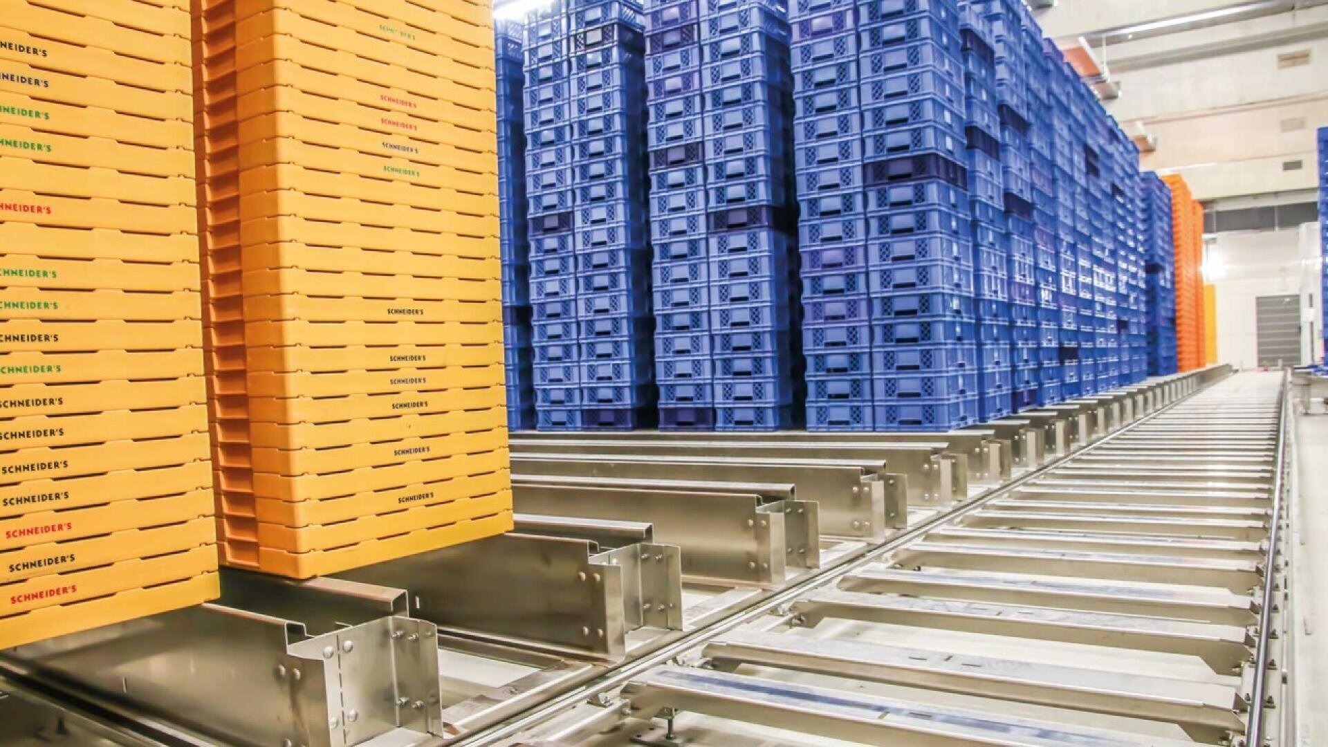 Tall stacks of yellow and blue plastic crates are arranged in a warehouse. The crates are organized on metal rails, likely part of an automated storage or conveyor system. The area is brightly lit and industrial.