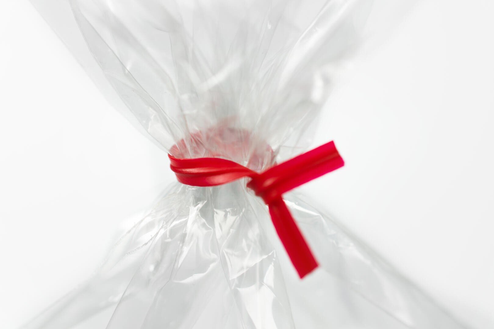 A close-up of clear cellophane gathered and tied with a red ribbon, creating a neat, decorative closure against a plain white background.