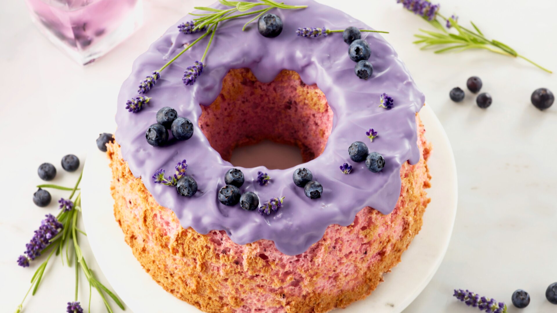 A round bundt cake with purple icing, topped with fresh blueberries and lavender sprigs, sits on a white marble platter. More blueberries, lavender, and two glasses of purple drink are on the marble surface.