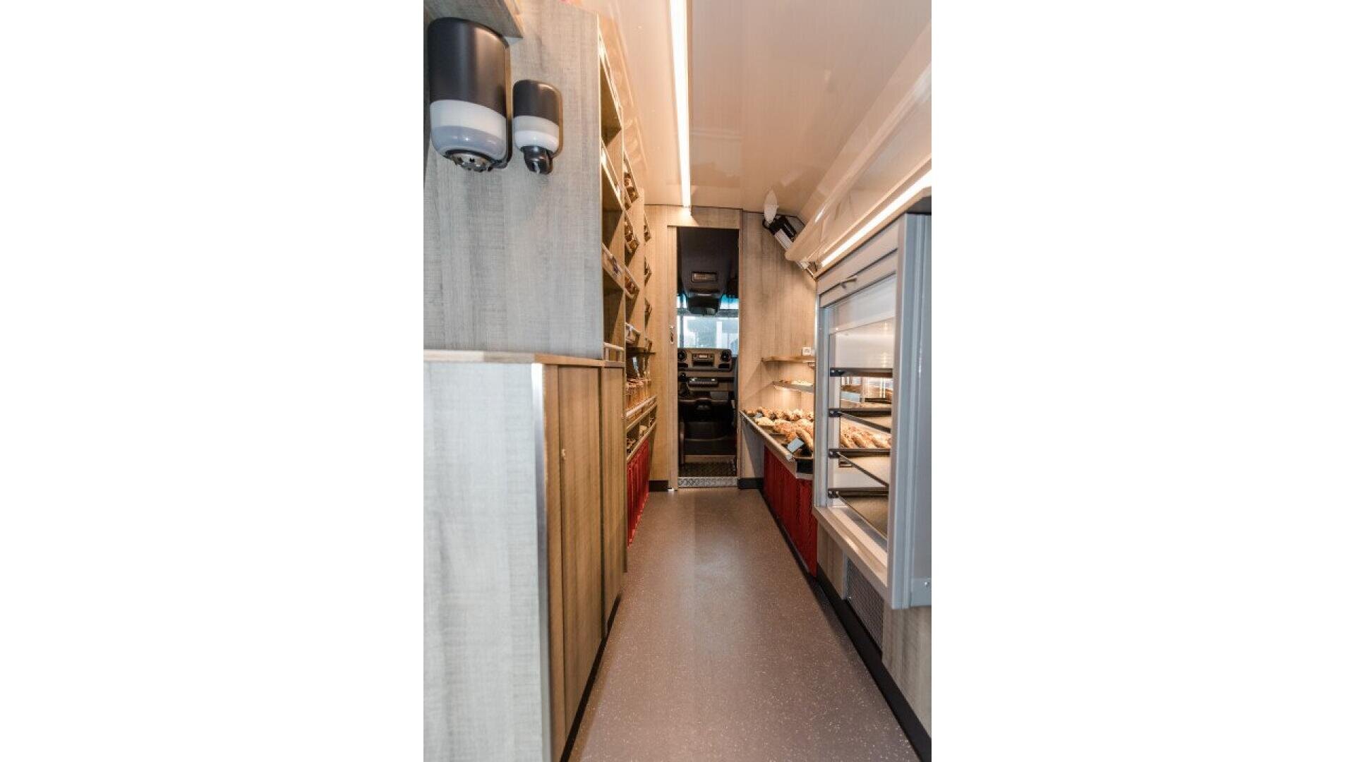 Interior of a narrow food truck or mobile kitchen, with shelves and cabinets on the left, refrigerated display cases on the right, and kitchen equipment visible at the far end.