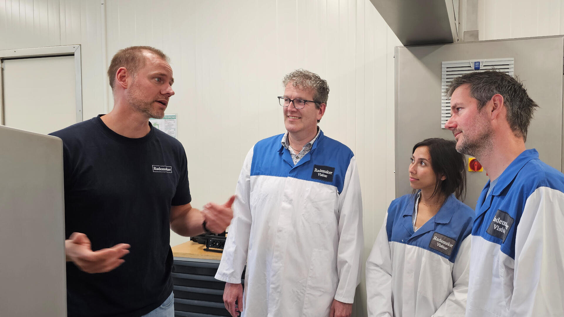 Four people stand in a laboratory, three in white and blue lab coats listening to a person in a black shirt who is speaking and gesturing with his hands. They appear to be having a discussion.
