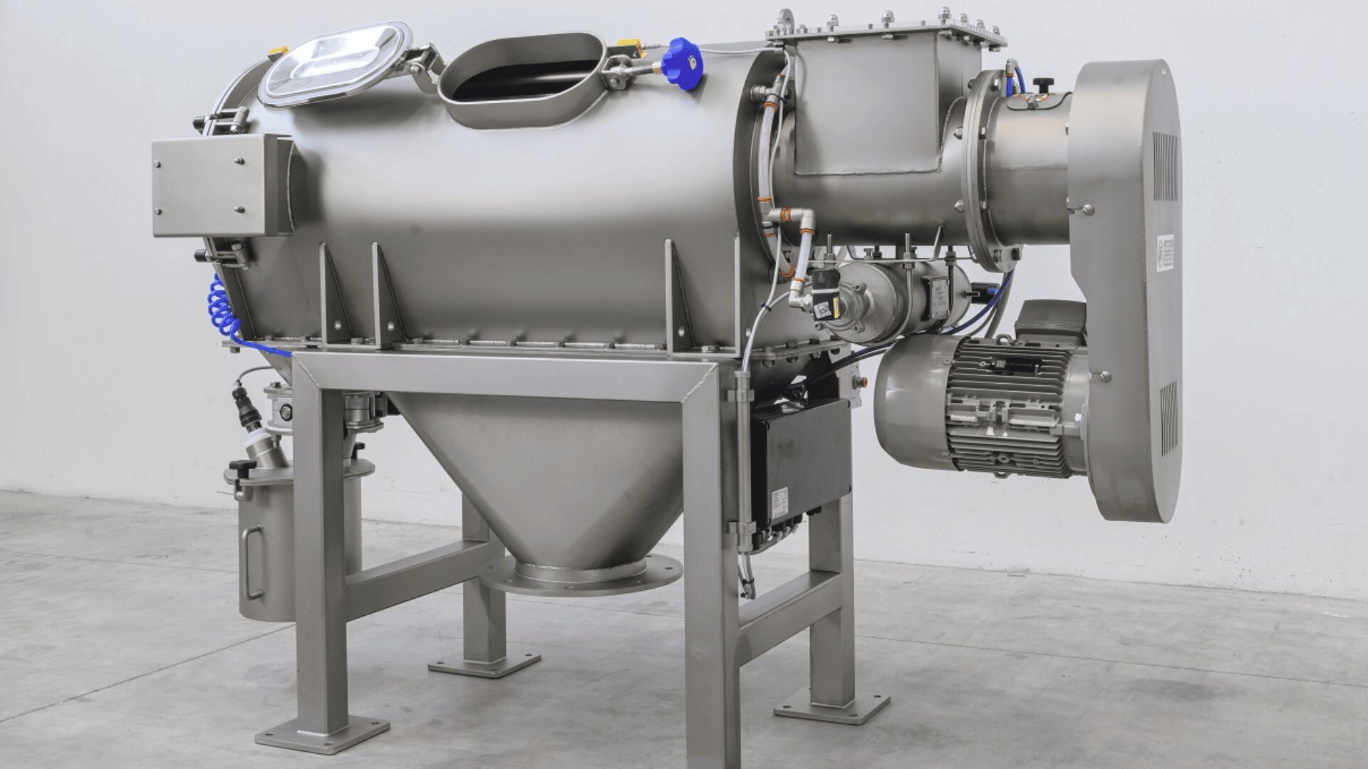 A large industrial stainless steel machine with a cylindrical body, conical bottom, and multiple attached components, standing on four legs in a clean, empty room with a concrete floor.