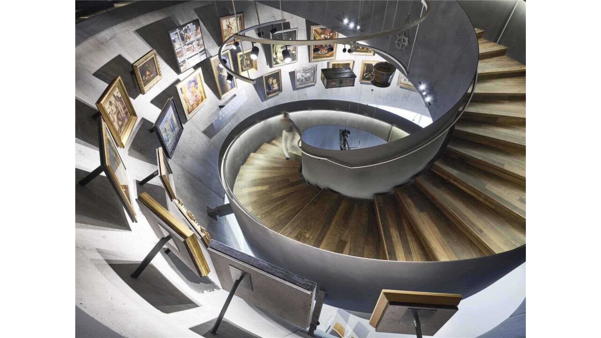 A person descends a modern, spiral wooden staircase surrounded by curved walls displaying framed artwork in a well-lit gallery space.