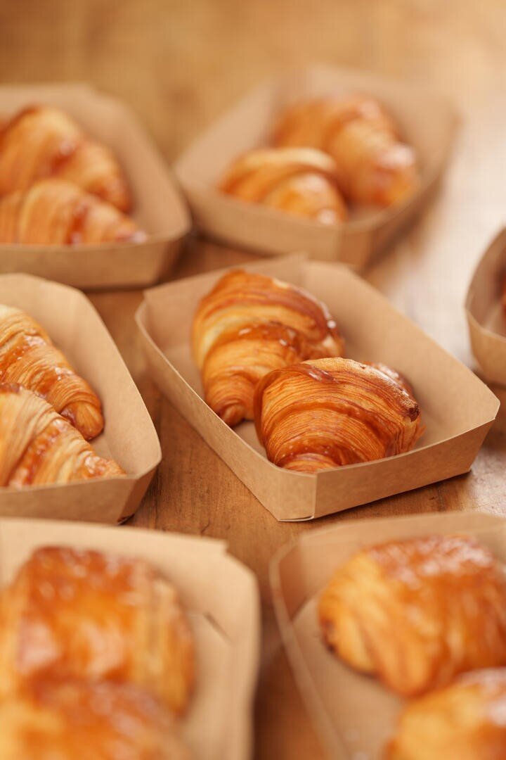 Several golden-brown croissants are arranged in small rectangular brown paper trays on a wooden surface, with a soft focus on the background.