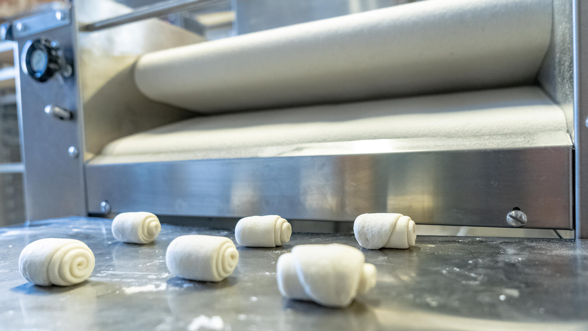 Several small rolled pieces of dough sit on a metal countertop in front of a stainless steel dough sheeter machine in a bakery setting.