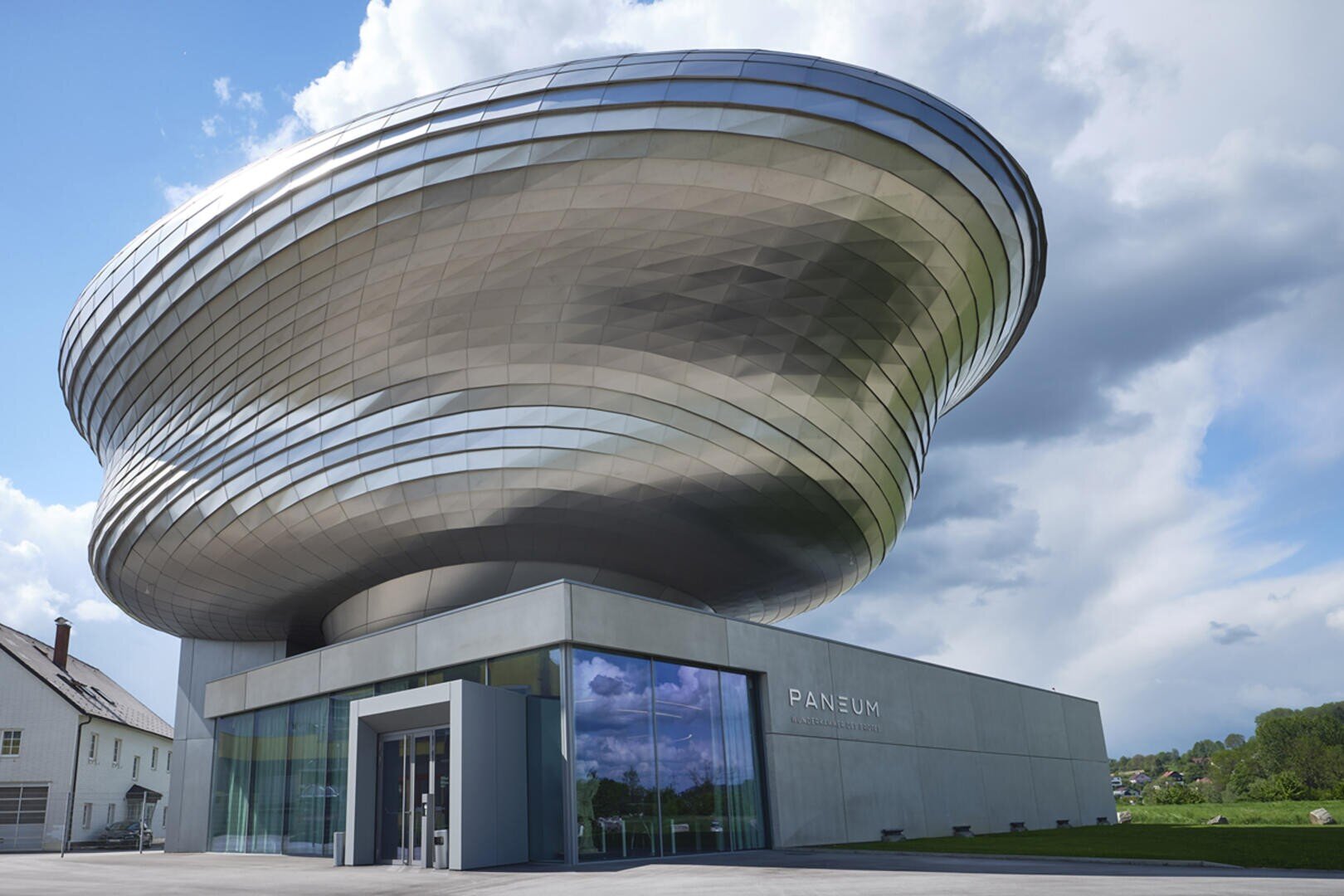 Ein modernes Gebäude mit einer großen, metallischen, spiralförmigen Struktur auf einem Sockel aus Glas und Beton, vor einem teilweise bewölkten Himmel. Auf der Seite des Gebäudes steht PANEUM geschrieben.