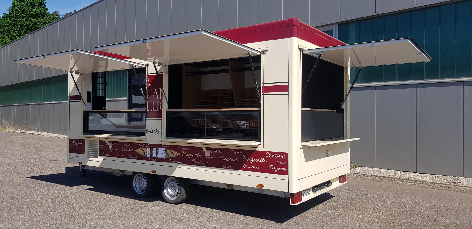 Ein beiger und roter Lebensmittelanhänger mit großen, offenen Servierfenstern ist auf einer gepflasterten Fläche neben einem modernen Industriegebäude geparkt. Der Anhänger ist mit lebensmittelbezogenen Wörtern beschriftet und hat im Inneren Regale.
