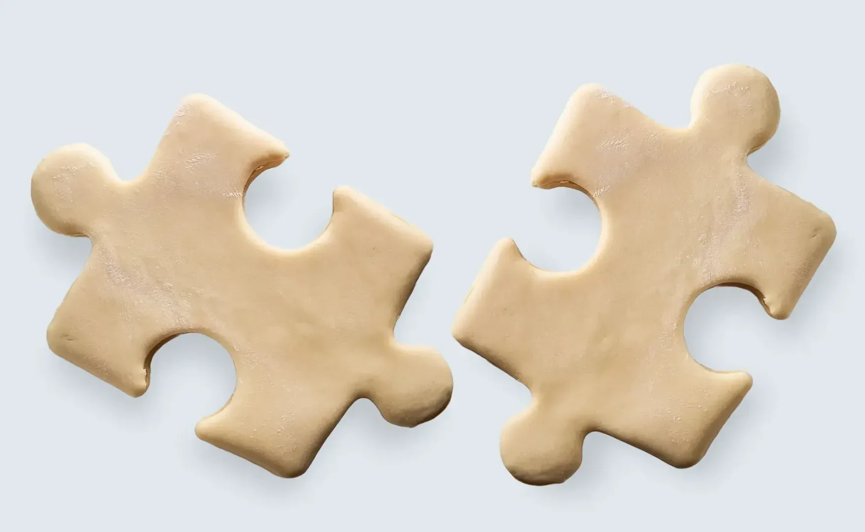 Two beige-colored puzzle pieces made of dough are placed side by side on a light background, resembling cookie dough cut into puzzle shapes.