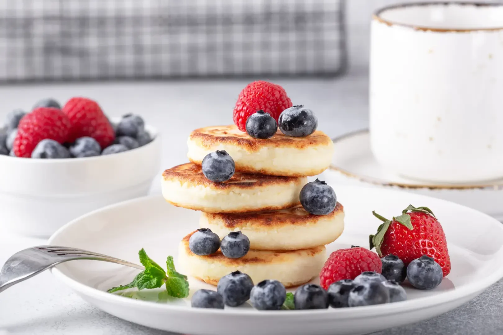 A stack of three pancakes topped with blueberries and raspberries on a white plate, with a fork, mint leaves, and more berries. In the background, a bowl of berries and a white mug are visible.