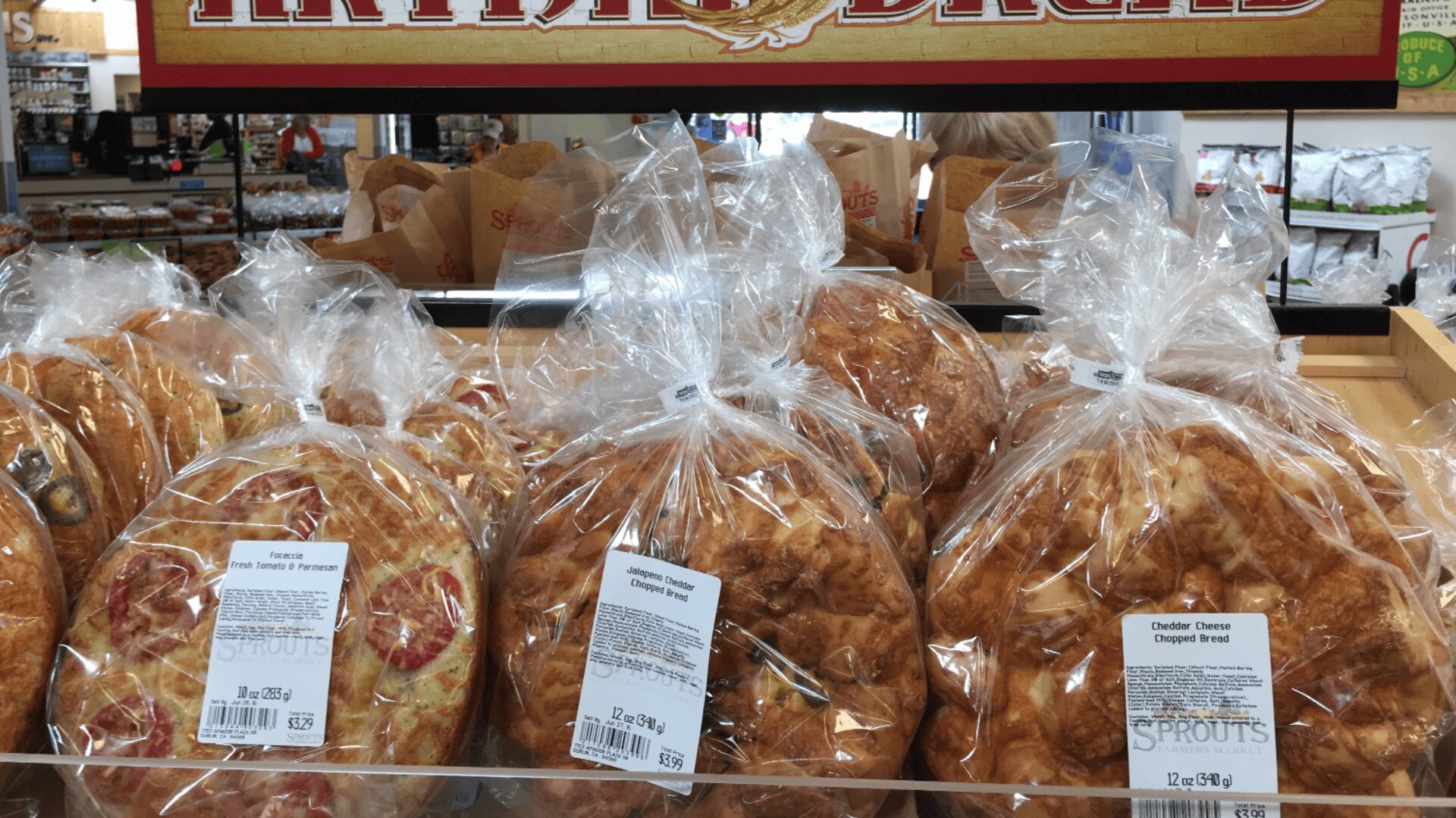 Mehrere in durchsichtiges Plastik eingewickelte Brote liegen in einem Regal aus. Auf dem oberen Etikett steht ARTISAN BREAD, und die sichtbaren Etiketten weisen auf die Käsesorten Tomate, Parmesan und Cheddar hin.