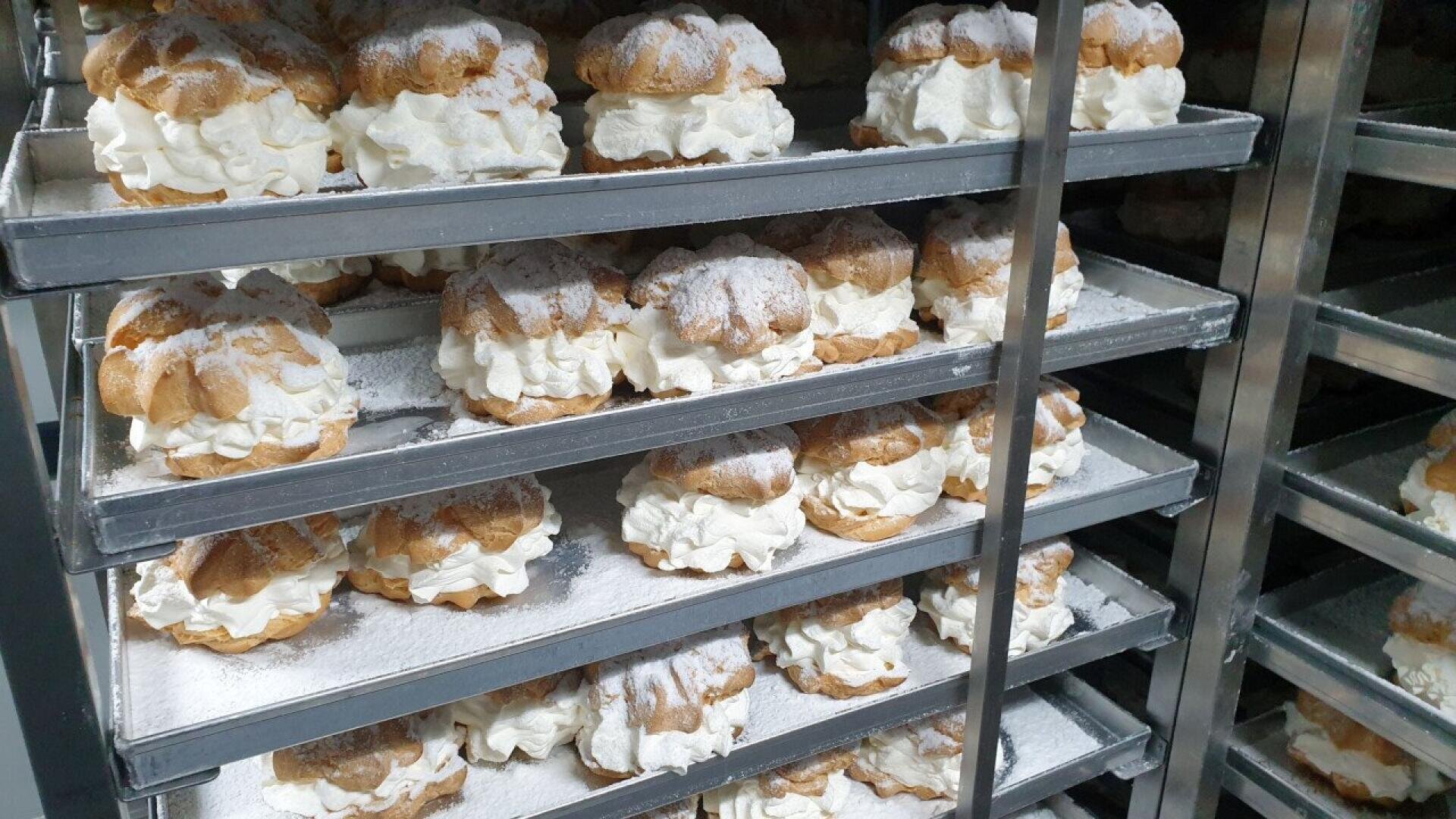 Trays filled with rows of large cream puffs, generously dusted with powdered sugar, stacked on metal rack shelves in a bakery setting.