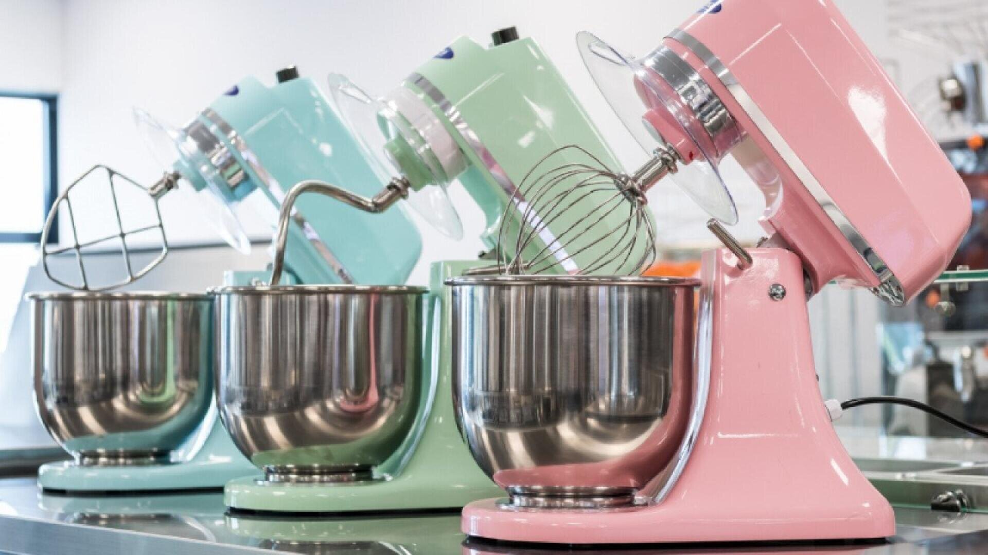 Drei Standmixer in Pastellfarben - rosa, mintgrün und hellblau - sind auf einer Arbeitsplatte aufgereiht. Jeder Mixer hat eine große Edelstahlschüssel und verschiedene Aufsätze. Im Hintergrund ist eine helle Küche zu sehen.