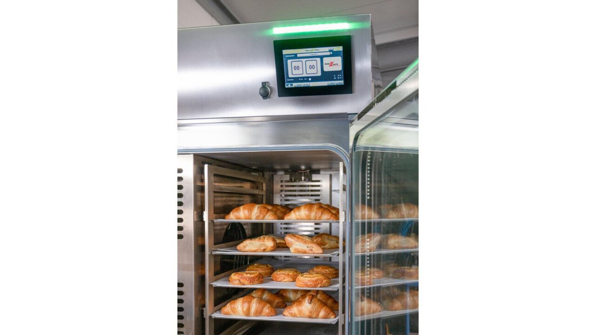Open industrial oven with trays of freshly baked croissants and pastries inside. A digital control panel with a display screen is mounted above the oven, showing settings and indicators.