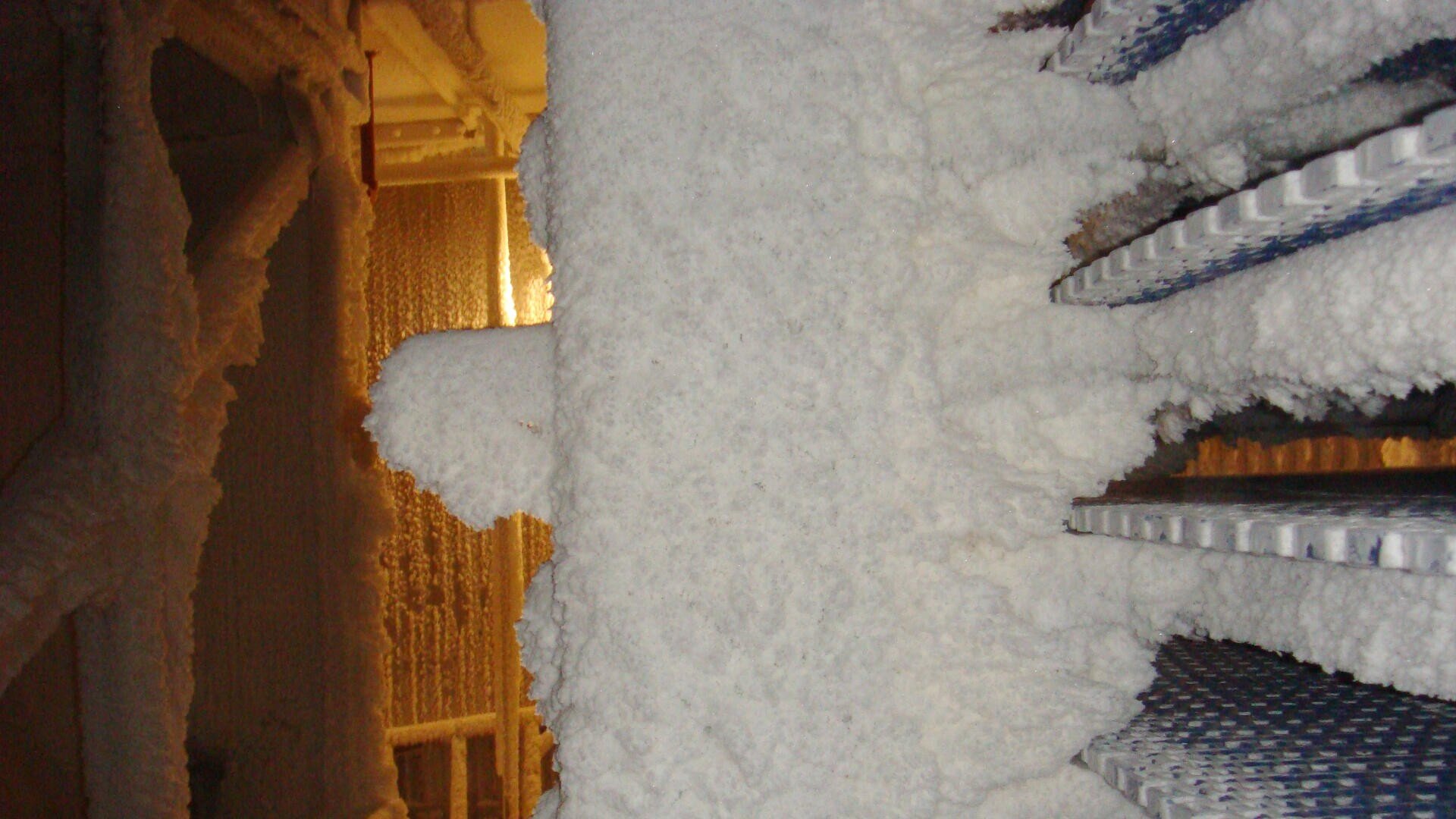 A thick layer of frost and ice covers industrial equipment and pipes, creating a textured, white surface. Blue metal shelves are partially visible on the right, and a faint, warm light illuminates the background.