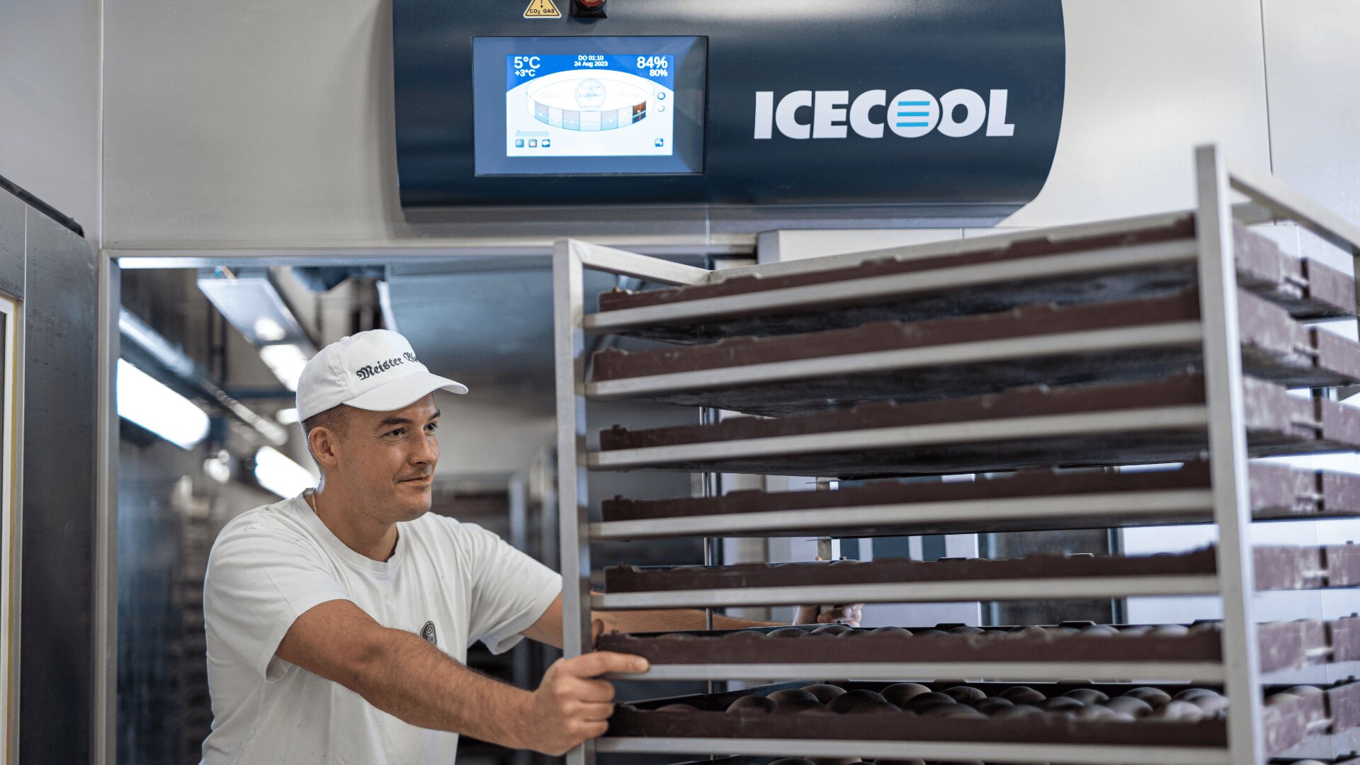 Ein Arbeiter in weißer Uniform und Mütze bewegt ein Regal mit Schalen, die mit Pralinen gefüllt sind, in einer Einrichtung. Im Hintergrund sind eine Maschine mit der Aufschrift ICECOOL und ein digitales Bedienfeld zu sehen.