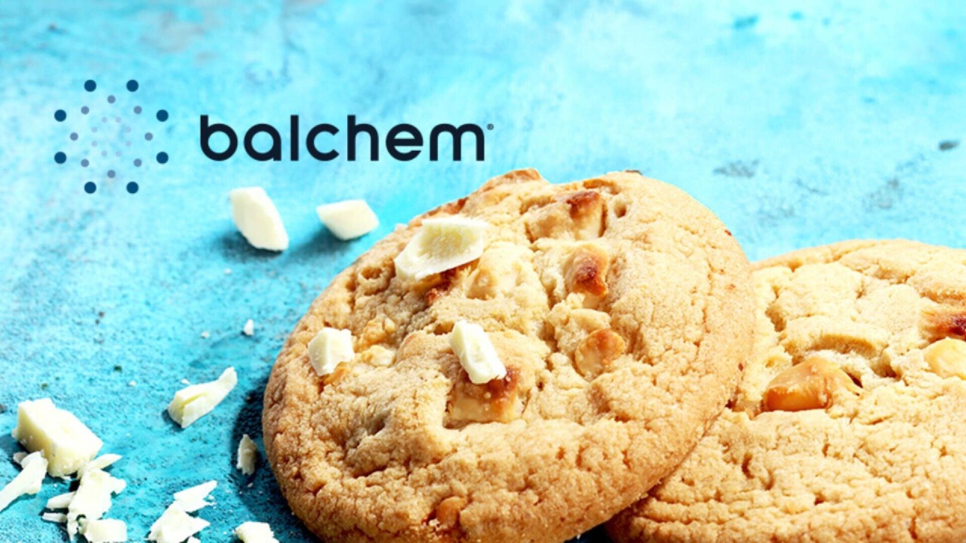 Zwei weiße Schokoladenkekse liegen auf einer blauen Fläche, neben verstreuten Stücken weißer Schokolade. Das Balchem-Logo erscheint in der oberen linken Ecke.