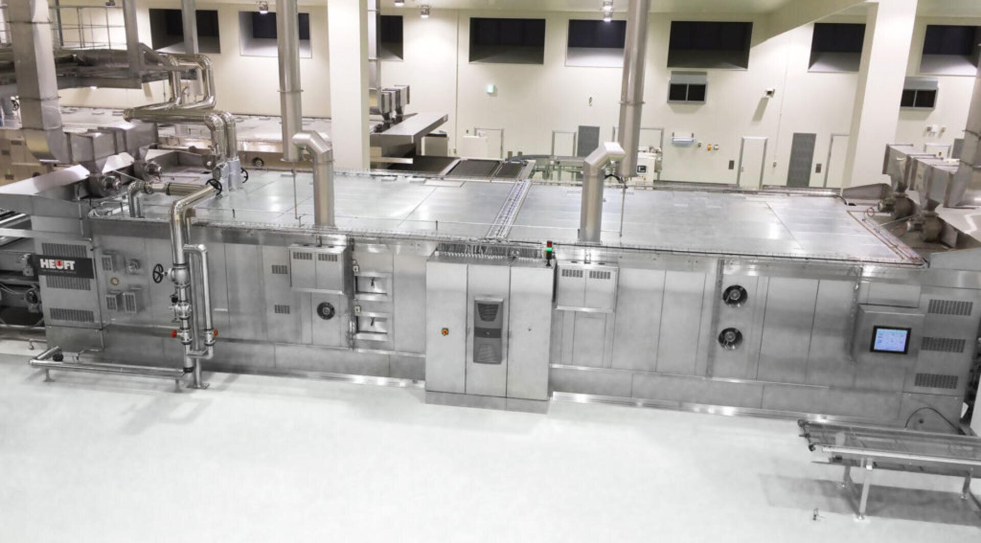 Eine große, industrielle Lebensmittelverarbeitungsmaschine mit Edelstahltafeln, Rohren, Schalttafeln und Bildschirmen in einer sauberen, hell erleuchteten Fabrikumgebung.