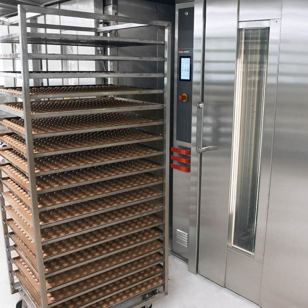 In einer sauberen, modernen Einrichtung steht ein großes Metallgestell, das mit Schalen mit gleichmäßig verteilten braunen Eiern gefüllt ist, neben einem Industrieofen oder Brutkasten aus Edelstahl.