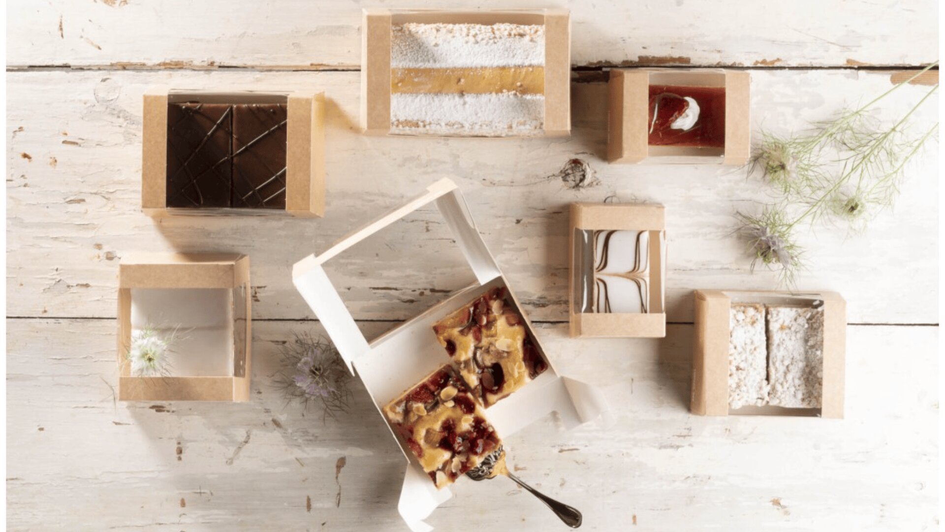 Assorted pastries in small brown cardboard boxes are arranged on a rustic white wooden table, with some delicate flowers scattered around and one box open to reveal two pieces of fruit and nut bars.
