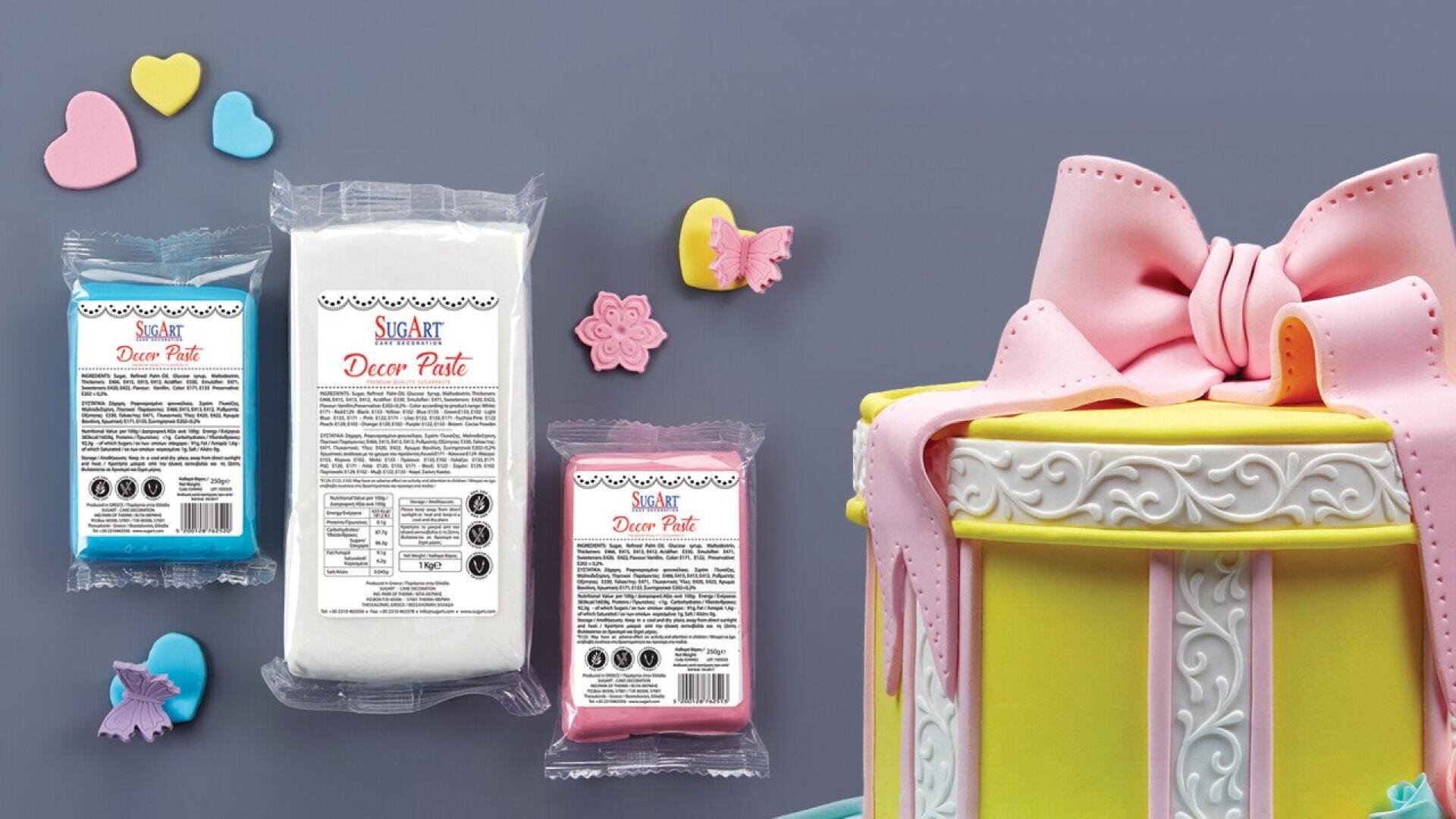 Drei Päckchen Sugar Art Decor Paste (blau, weiß, rosa) werden mit pastellfarbenen Fondantformen und einer dekorierten Torte mit einer großen rosa Schleife, gelben Verkleidungen und weißem Zuckerguss auf grauem Hintergrund präsentiert.