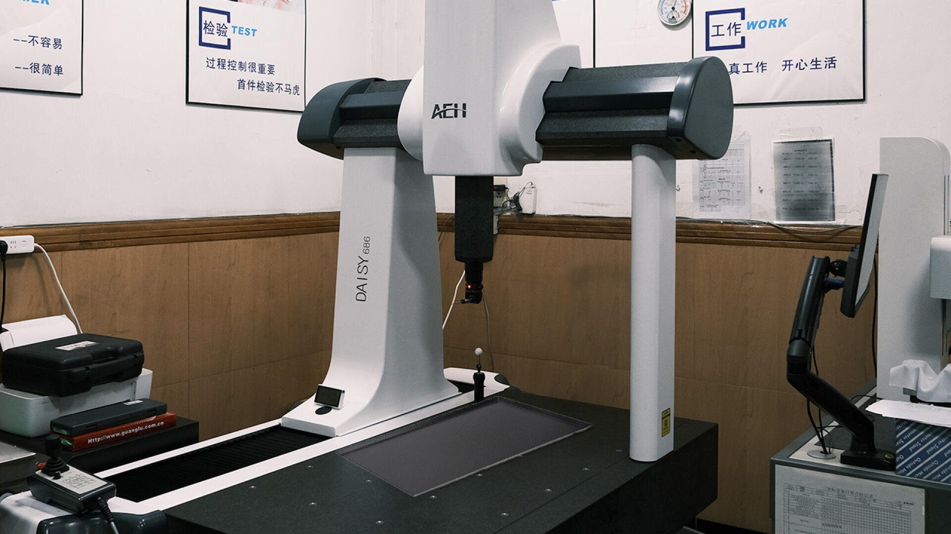 A coordinate measuring machine labeled DAISY stands in a lab, surrounded by office equipment and Chinese-language posters on the walls, with a computer monitor nearby.
