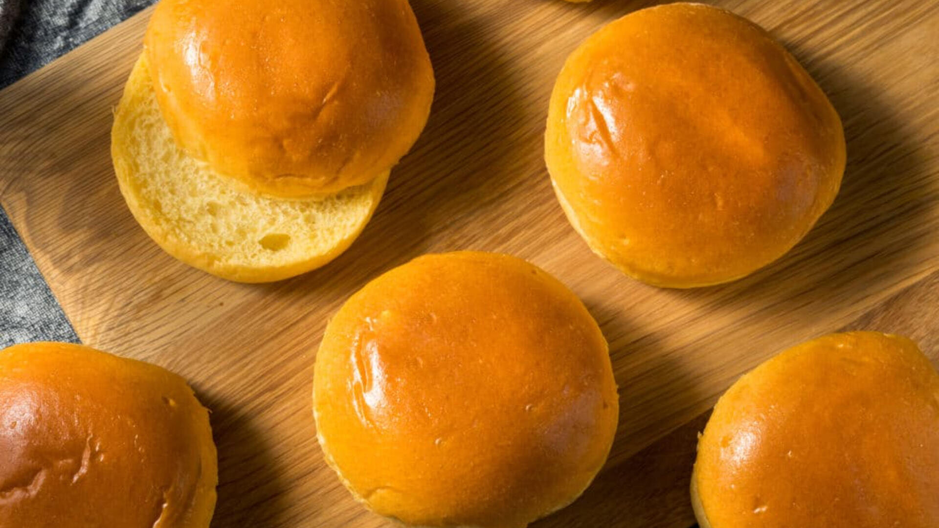 Mehrere glänzende, goldbraune Brioche-Brötchen liegen auf einem Schneidebrett aus Holz, wobei ein Brötchen aufgeschnitten ist und sein weiches, luftiges Inneres preisgibt.