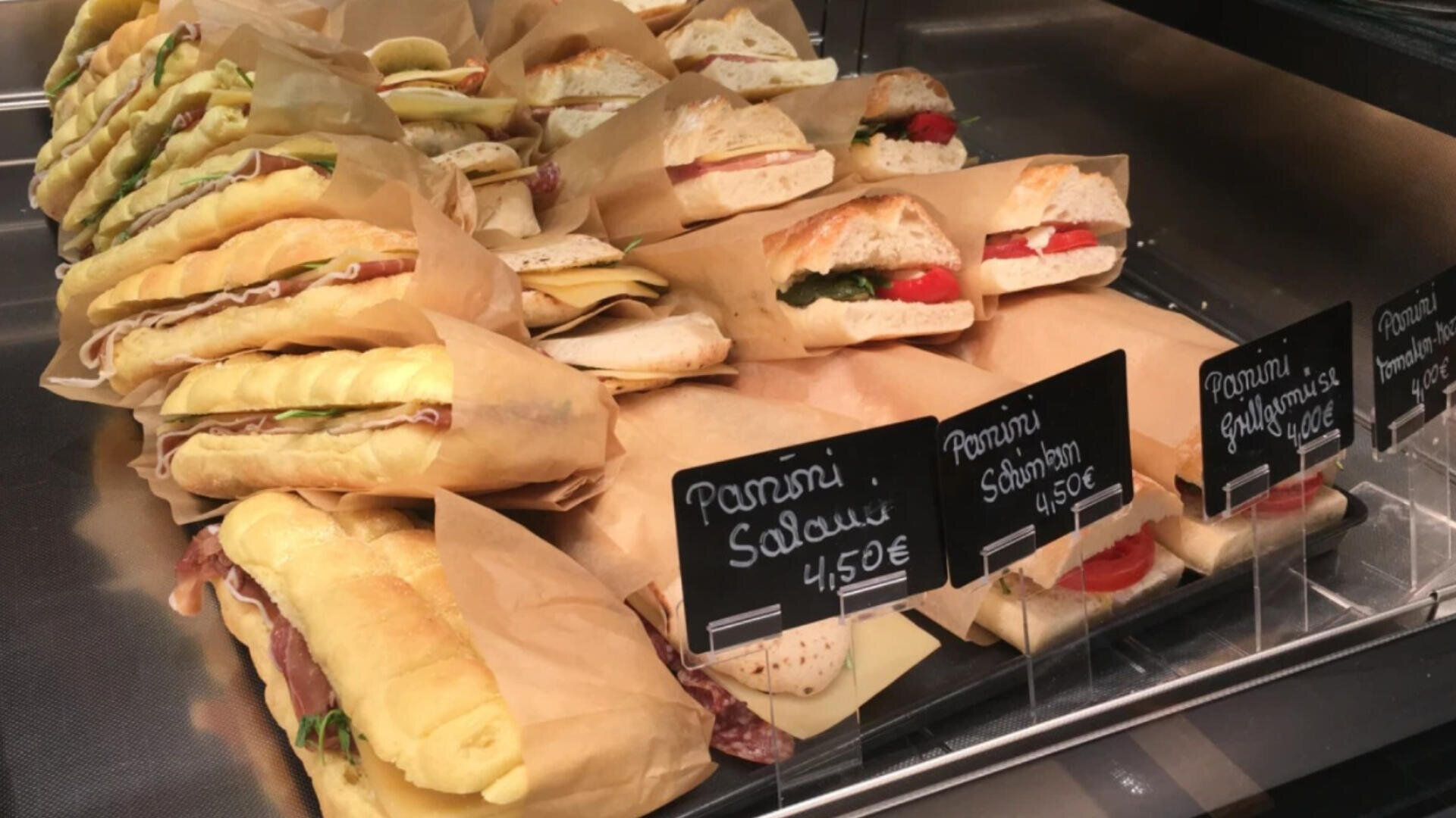 A display case shows various panini sandwiches wrapped in brown paper, each labeled with small black signs listing their names and prices in euros. The sandwiches are filled with meats, cheese, and vegetables.