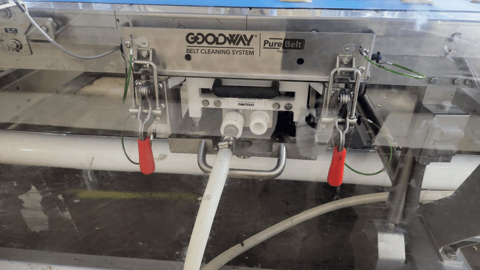 A Goodway PureBelt belt cleaning system with hoses attached and red-handled clamps, mounted on a conveyor belt in an industrial setting.