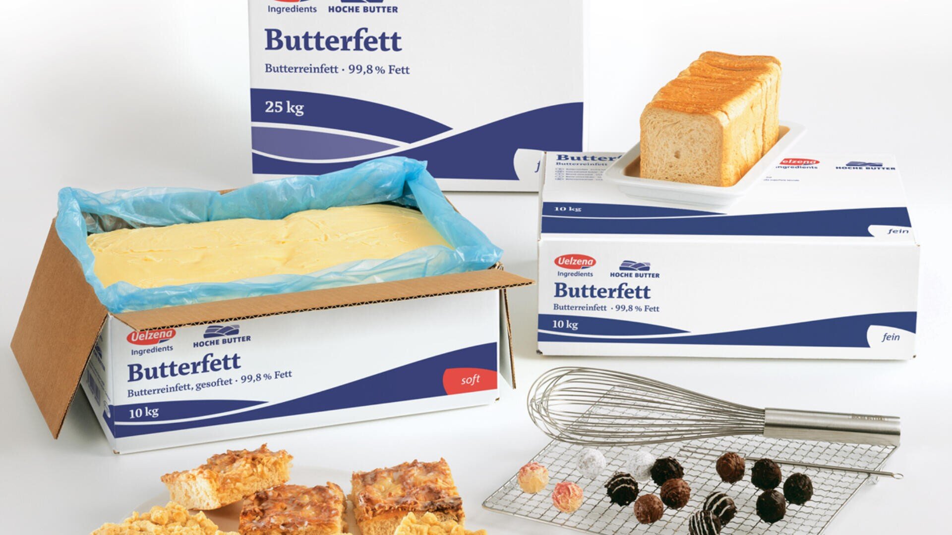 Auf einer weißen Fläche stehen Kartons mit der Aufschrift Butterfett, in denen ein großer Butterblock, ein Laib Brot, ein Schneebesen, Gebäck und Backzutaten angeordnet sind.