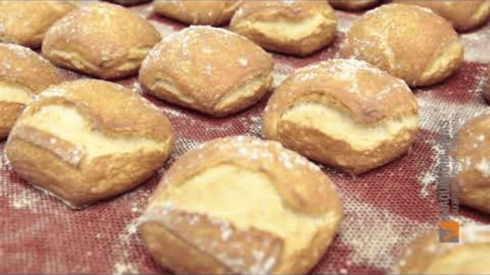 Rows of golden brown, freshly baked rolls dusted with flour are arranged on a red baking mat. The rolls have a cracked, rustic crust and a soft texture.