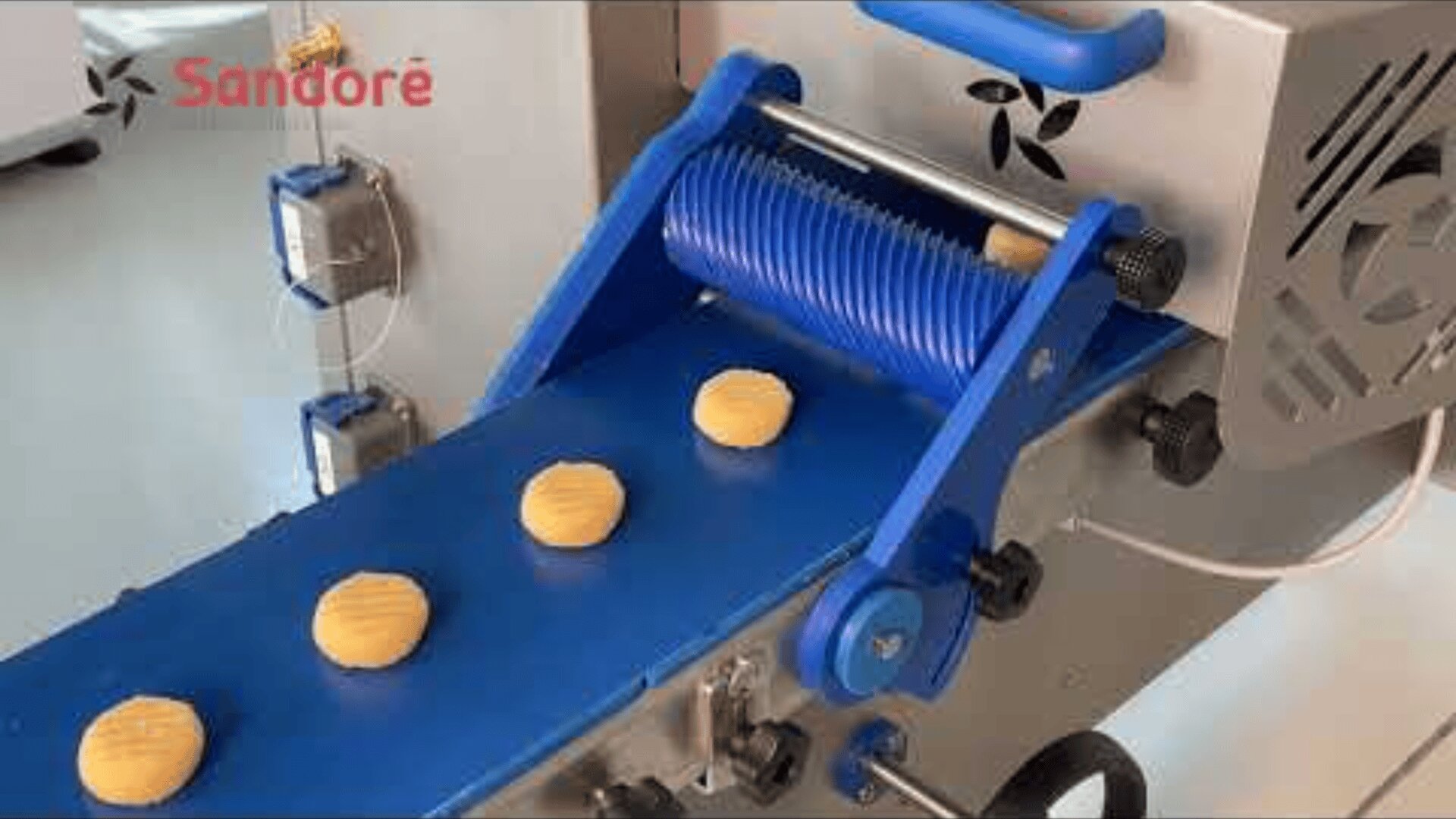 A close-up of a blue conveyor belt machine forming evenly spaced dough rounds, with the brand name “Sandoré” visible in the top left corner.